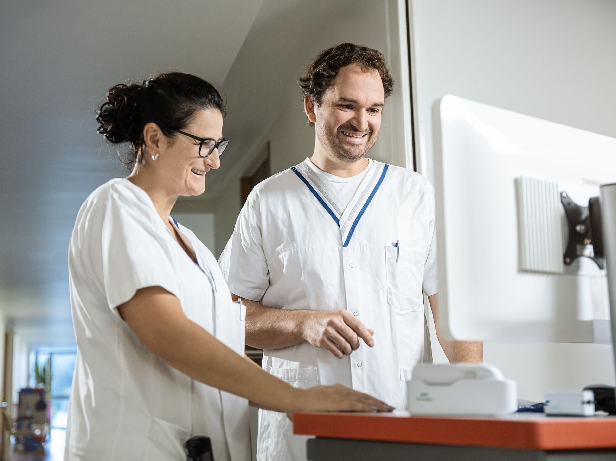 Zwei Pflegekräfte im Gespräch vor einem Computerbildschirm, beide lächeln und wirken konzentriert.