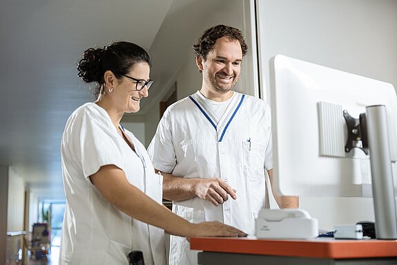 Zwei Pflegekräfte im Gespräch vor einem Computerbildschirm, beide lächeln und wirken konzentriert.