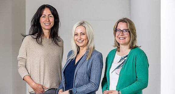Das Bild zeigt drei Frauen, die in die Kamera lächeln.