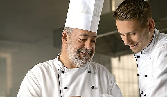 Ein alter Koch zeigt einem jungen Koch etwas.