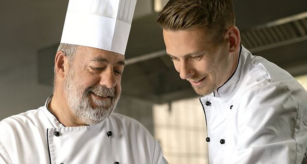 Ein alter Koch zeigt einem jungen Koch etwas.