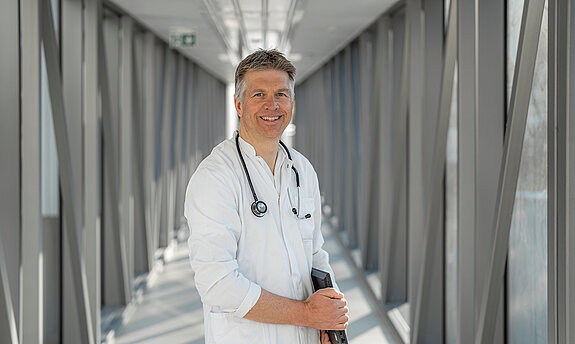 Ein lächelnder Arzt steht mit einer Mappe im Gang.