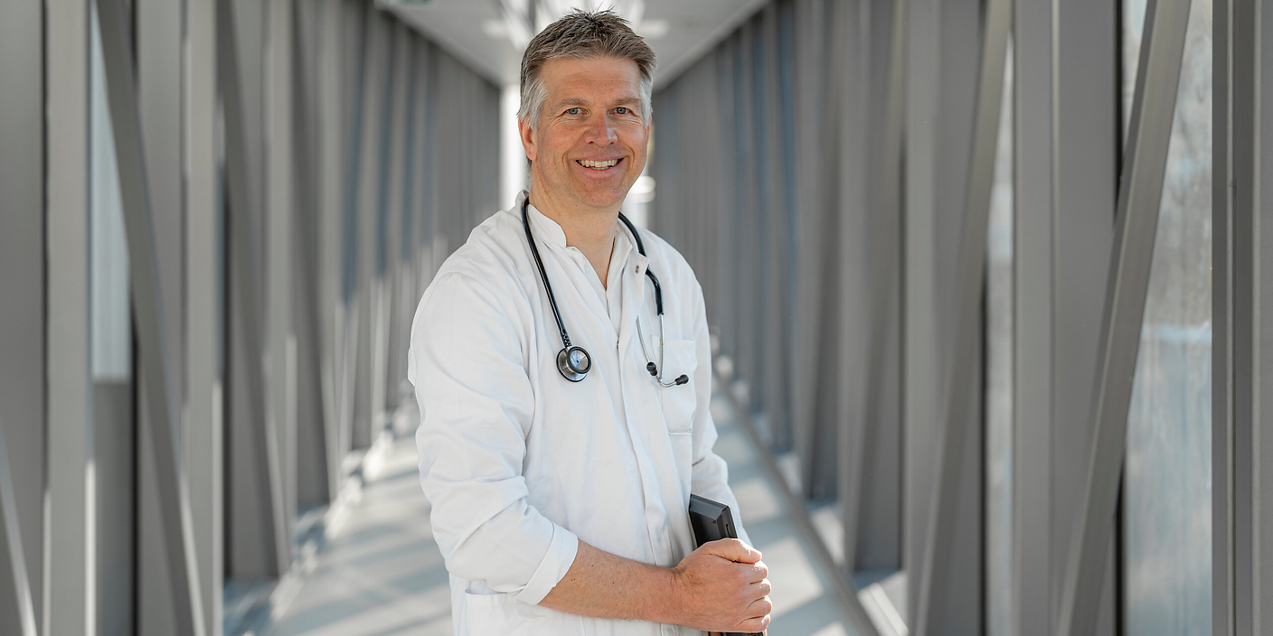 Ein lächelnder Arzt steht mit einer Mappe im Gang.