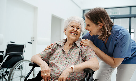Eine ältere Patientin im Rollstuhl wird freundlich von einer Pflegerin betreut.