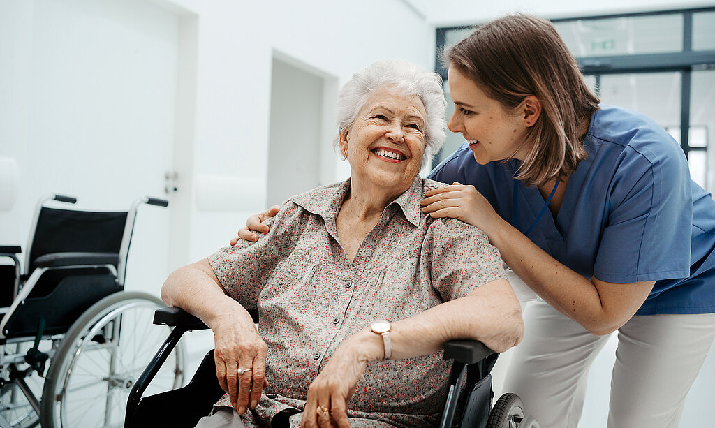 Eine ältere Patientin im Rollstuhl wird freundlich von einer Pflegerin betreut.