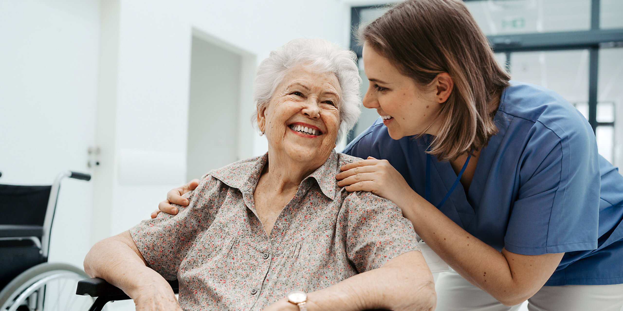 Eine ältere Patientin im Rollstuhl wird freundlich von einer Pflegerin betreut.