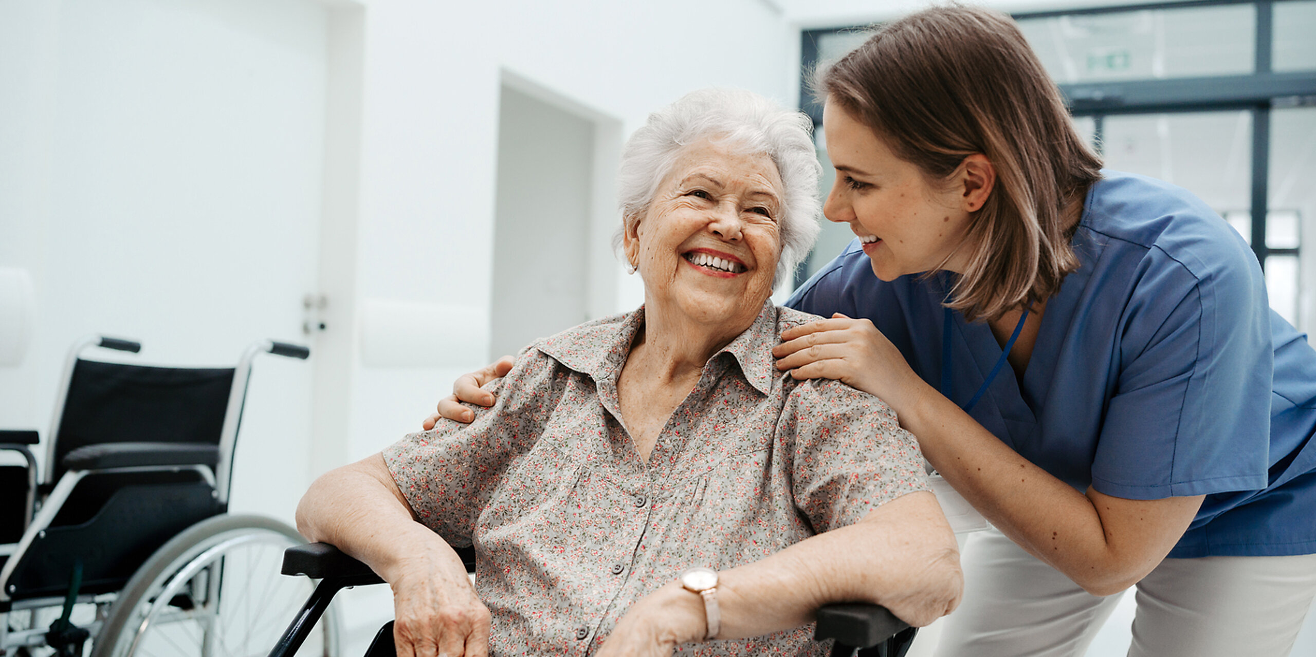 Eine ältere Patientin im Rollstuhl wird freundlich von einer Pflegerin betreut.