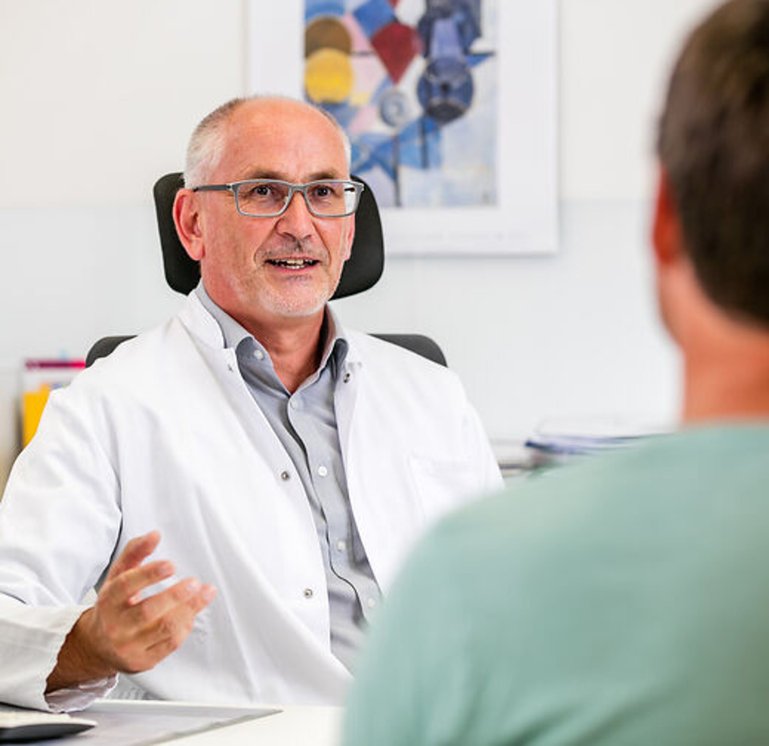 Ein Arzt unterhält sich freundlich mit einem Pateinten am Schreibtisch.