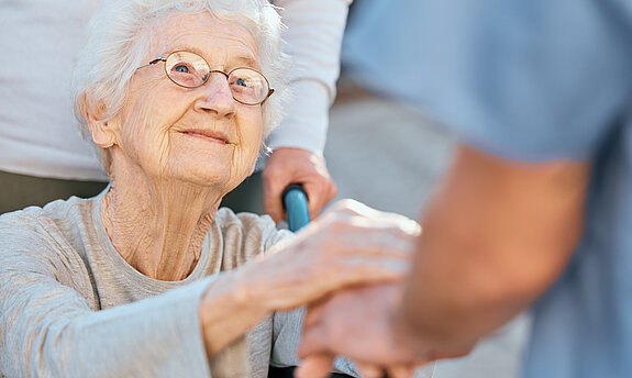Eine Seniorin im Rollstuhl hält dankbar die Hände einer Pflegekraft.