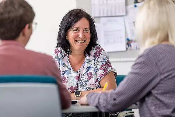 Eine Mitarbeiterin unterhält sich mit zwei Patienten am Schreibtisch.