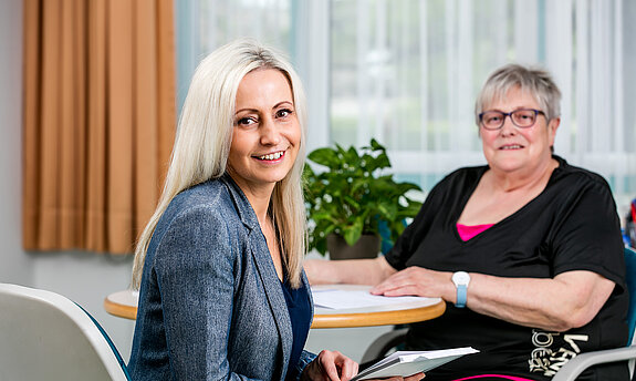Eine Mitarbeiterin aus dem Sozialdienst lächelt in die Kamera, eine Patientin sitzt - ebenfalls lächelnd - mit am Tisch.