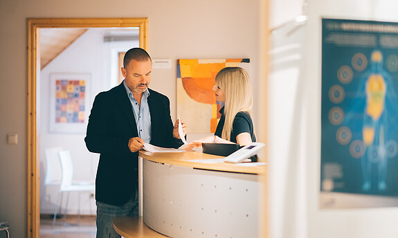 Dr. Köhler mit MFA an der Theke (Anmeldung). Beide sehen sich unterlagen an