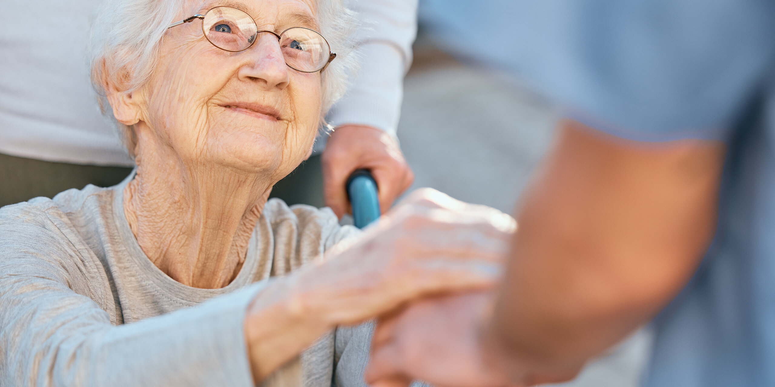 Eine Seniorin im Rollstuhl hält dankbar die Hände einer Pflegekraft.