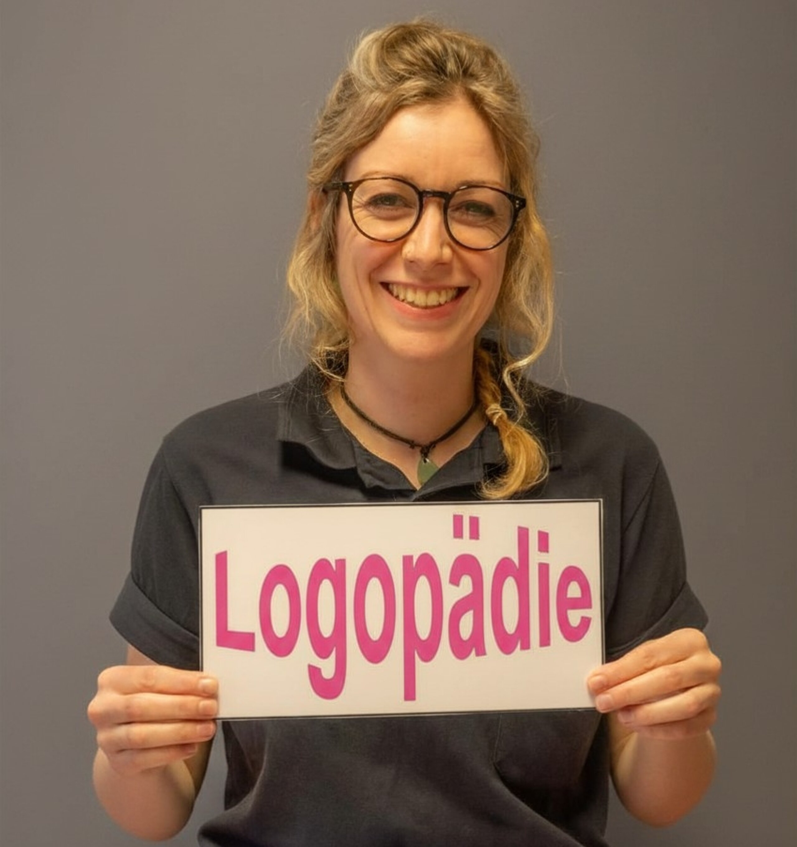 Freundlich lächelnde Logopädin hält ein Schild mit der Aufschrift ‚Logopädie‘ in der Hand.