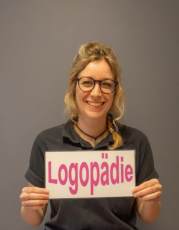 Freundlich lächelnde Logopädin hält ein Schild mit der Aufschrift ‚Logopädie‘ in der Hand.
