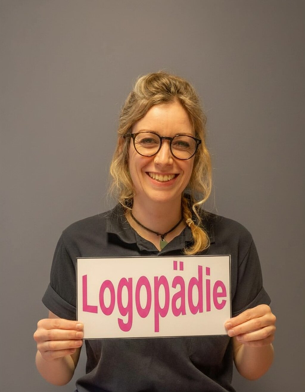 Freundlich lächelnde Logopädin hält ein Schild mit der Aufschrift ‚Logopädie‘ in der Hand.