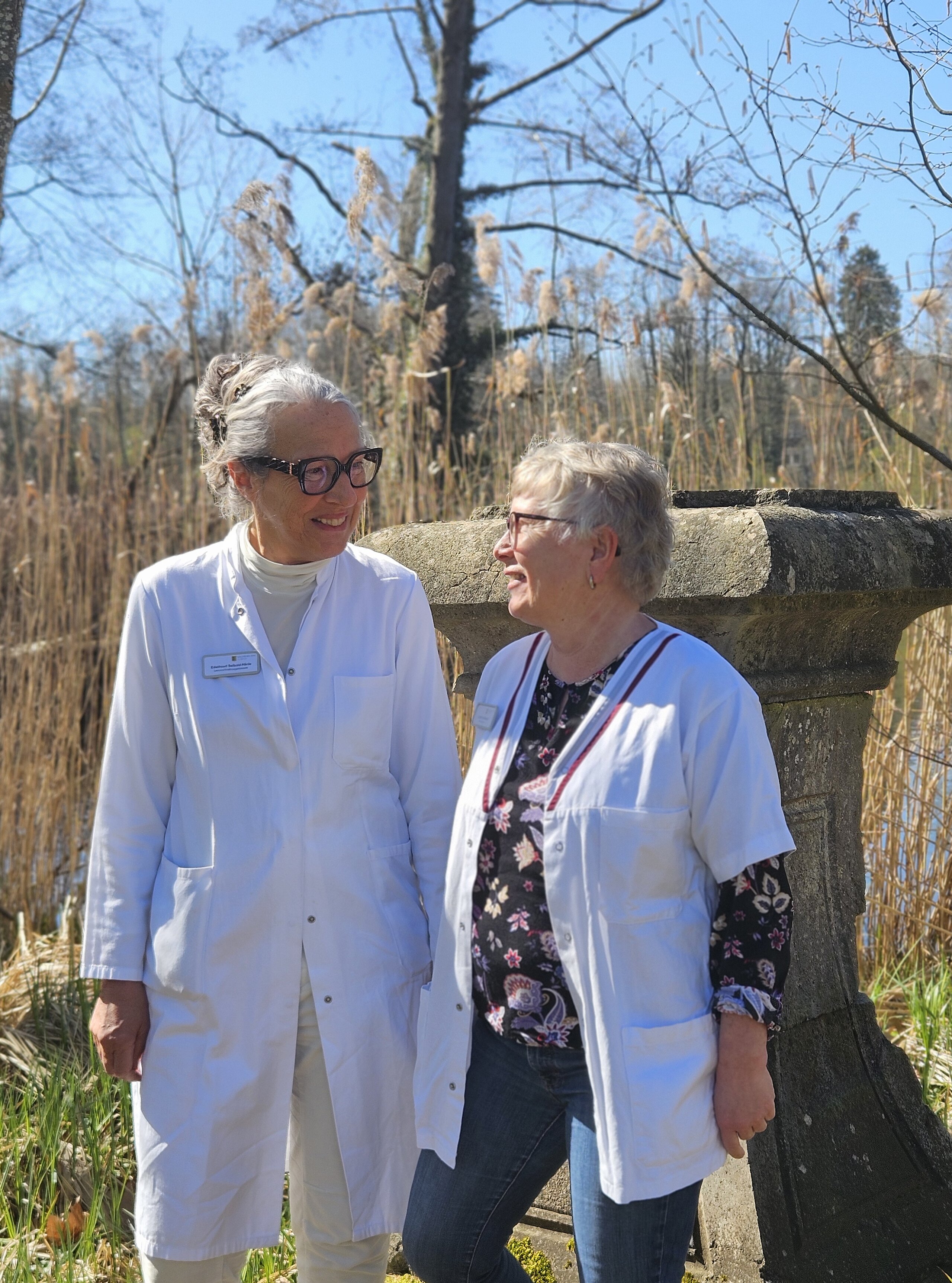 Zwei Mitarbeitende der Ernährungsberatung der Klinik im Hofgarten stehen in weißem Kittel lächelnd im Freien vor einem bewaldeten Hintergrund