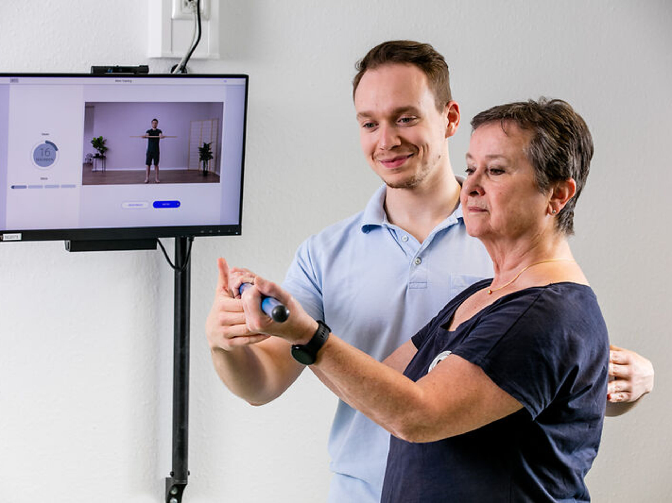 Ein Therapeut unterstützt eine Patientin in ihrem Training, welches sie anhand eines Trainingsvideos durchführt.