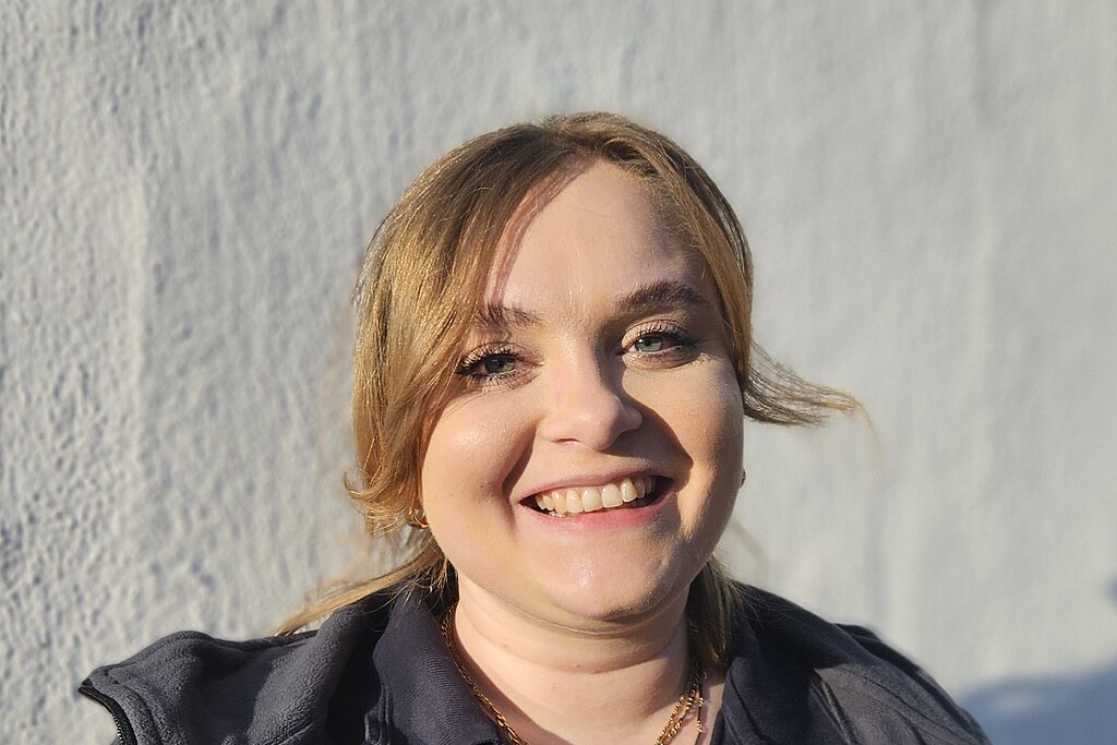 Eine junge Therapeutin der KIH steht vor einer weißen Wand und trägt Therapie- Arbeitsbekleidung und lächelt.