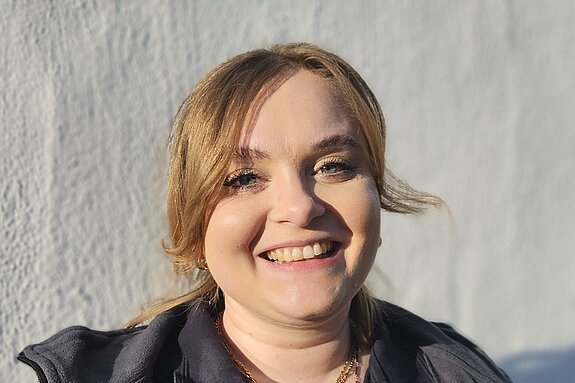 Eine junge Therapeutin der KIH steht vor einer weißen Wand und trägt Therapie- Arbeitsbekleidung und lächelt.