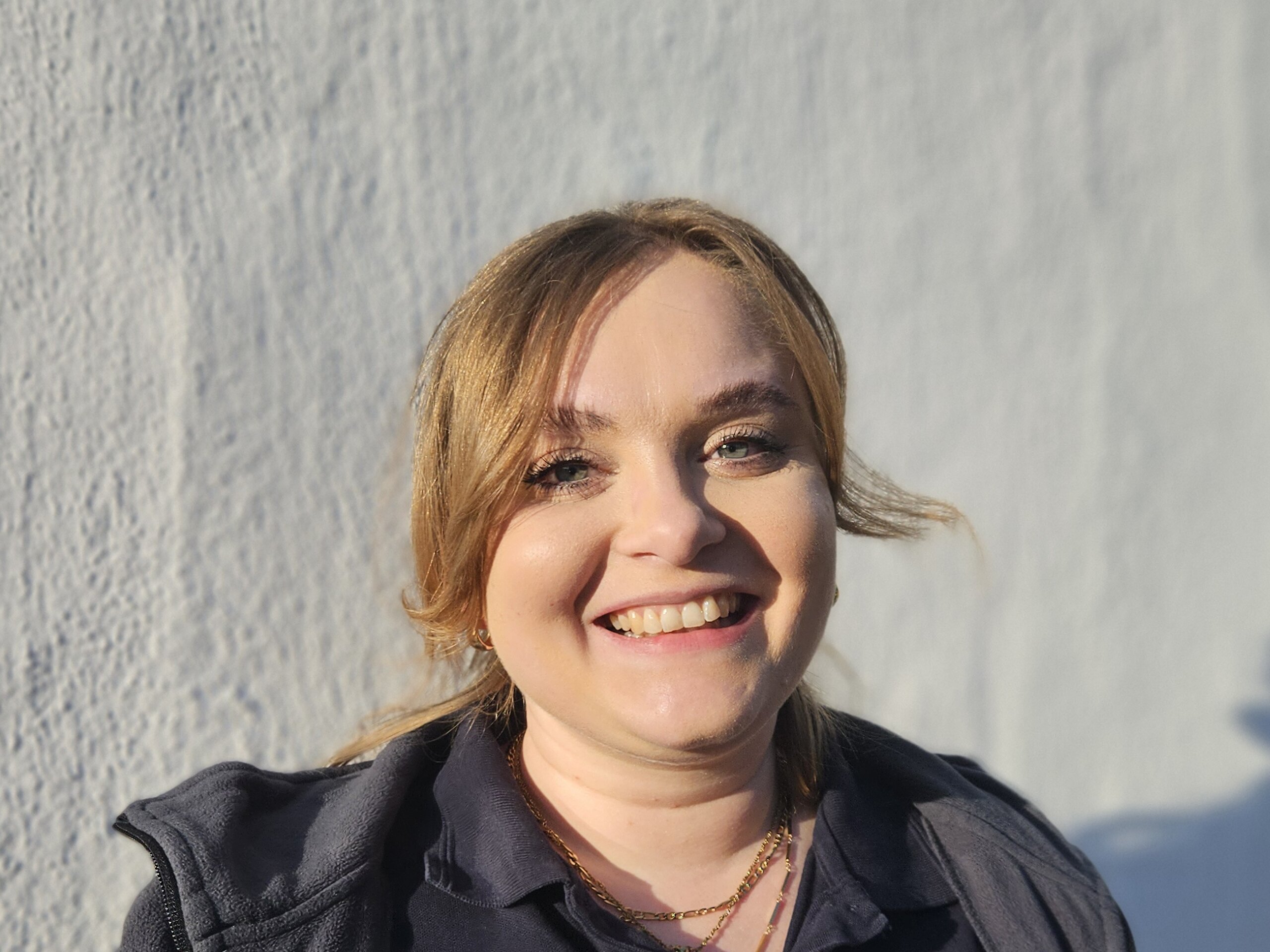 Eine junge Therapeutin der KIH steht vor einer weißen Wand und trägt Therapie- Arbeitsbekleidung und lächelt.