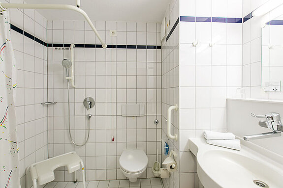 Das Bild zeigt ein Badezimmer mit Waschbecken, Toilette und Dusche mit Duschhocker.