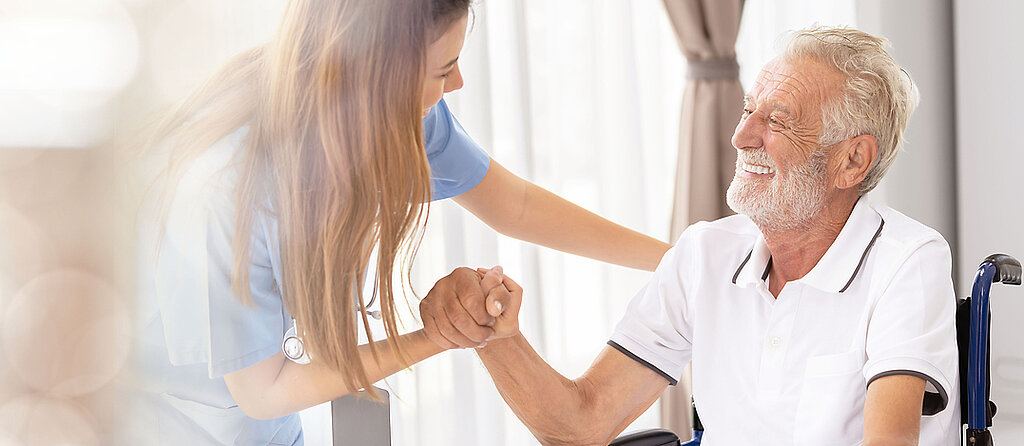 Ein älterer Patient im Rollstuhl wird freundlich von einer Pflegekraft angesprochen und betreut.