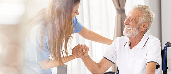 Ein älterer Patient im Rollstuhl wird freundlich von einer Pflegekraft angesprochen und betreut.