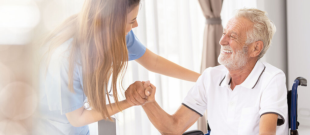 Ein älterer Patient im Rollstuhl wird freundlich von einer Pflegekraft angesprochen und betreut.