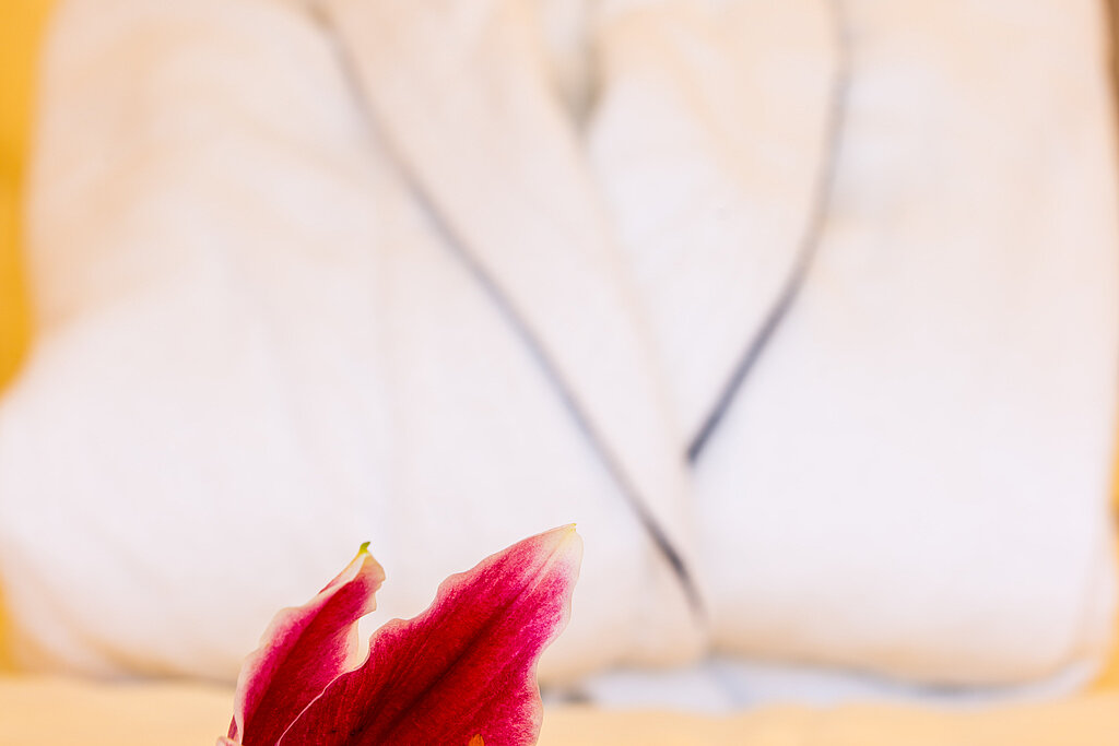 Ein weißer Bademantel liegt dekorativ auf einem Bett, im Vordergrund eine pinkfarbene Blume – Symbol für Wohlfühlatmosphäre und Entspannung.
