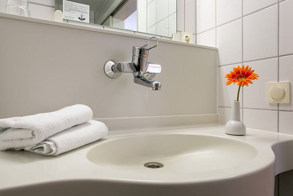 Das Bild zeigt ein Waschbecken, auf dem zwei weiße, gefaltete Handtücher liegen sowie eine Vase mit einer orangenen Blume steht.