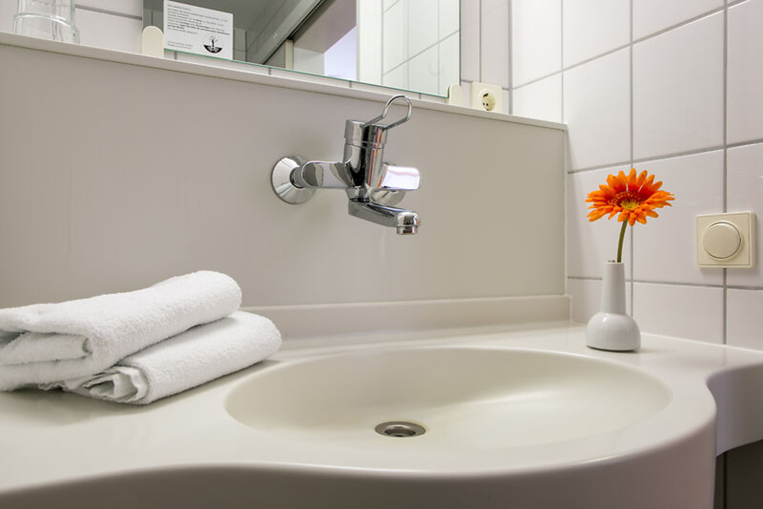 Das Bild zeigt ein Waschbecken, auf dem zwei weiße, gefaltete Handtücher liegen sowie eine Vase mit einer orangenen Blume steht.