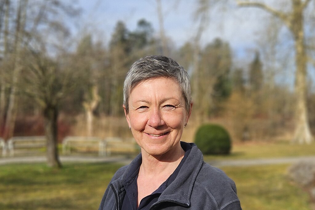 Eine Therapeutin steht im Park, trägt eine dunkle Jacke mit Bäume und Wiese im Hintergrund.