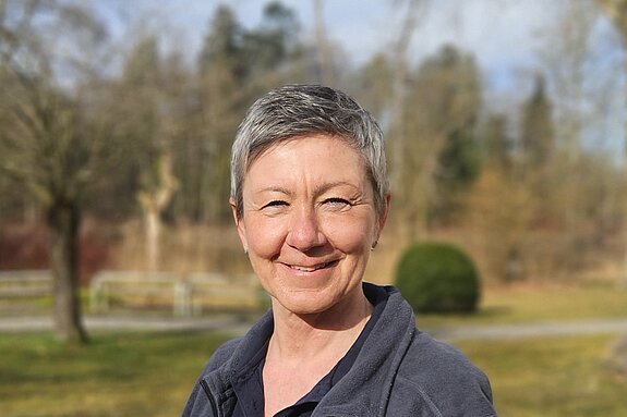 Eine Therapeutin steht im Park, trägt eine dunkle Jacke mit Bäume und Wiese im Hintergrund.