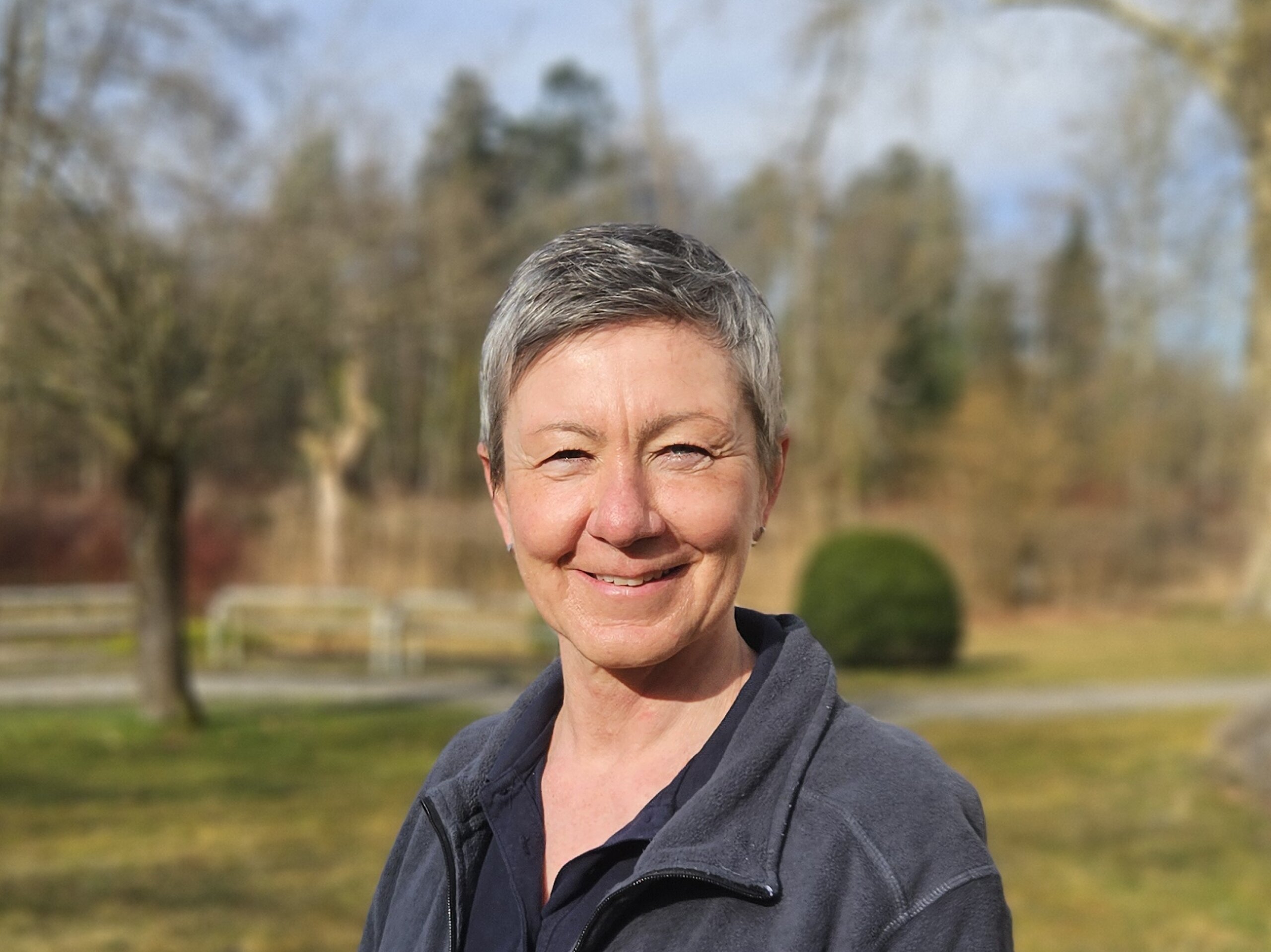 Eine Therapeutin steht im Park, trägt eine dunkle Jacke mit Bäume und Wiese im Hintergrund.