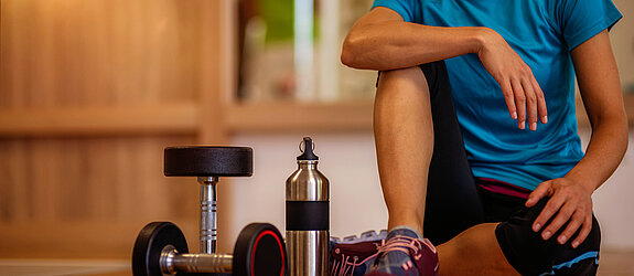 Eine Frau sitzt auf dem Boden eines Fitnessraums mit Hanteln und Trinkflasche.