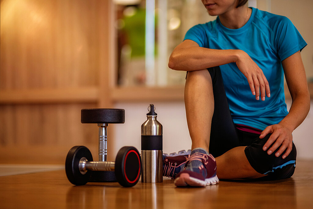 Eine Frau sitzt auf dem Boden eines Fitnessraums mit Hanteln und Trinkflasche.