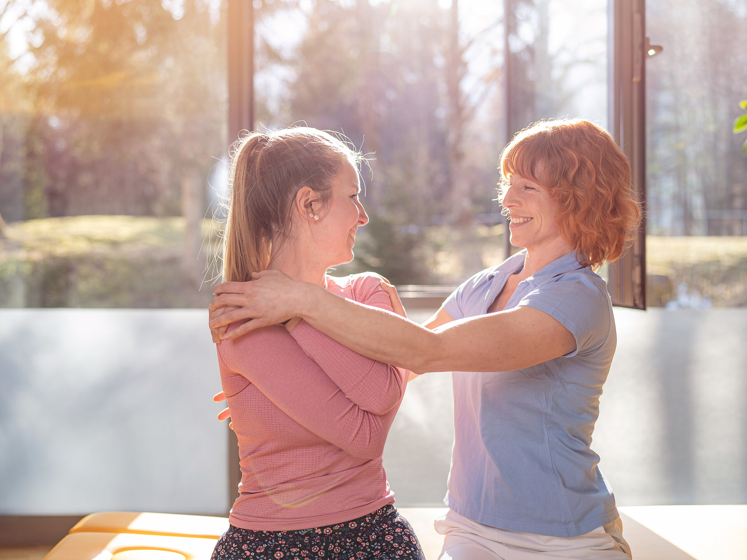 Zwei Frauen in einem hellen Raum führen eine physiotherapeutische Übung am Oberkörper durch und lächeln sich an