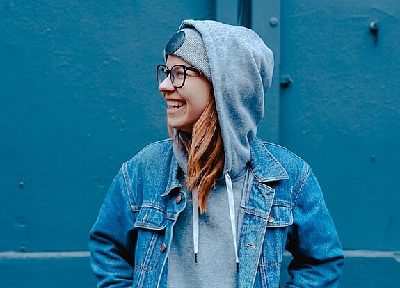 Eine Schülerin in Jeansjacke steht lachend vor einer blauen Wand.