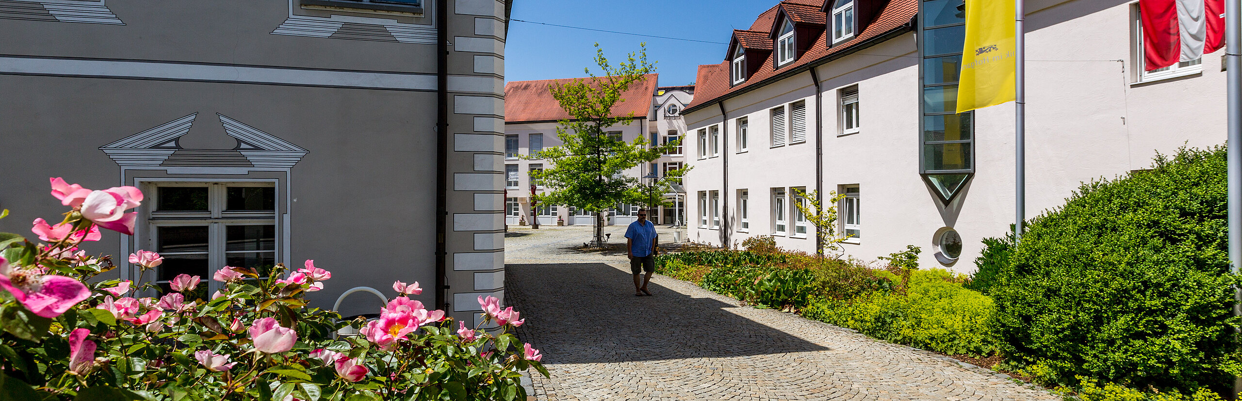 Zugang zur Klinik im Hofgarten mit gepflastertem Weg, Blumenbeeten und Gebäuden mit Fahnen an der Fassade
