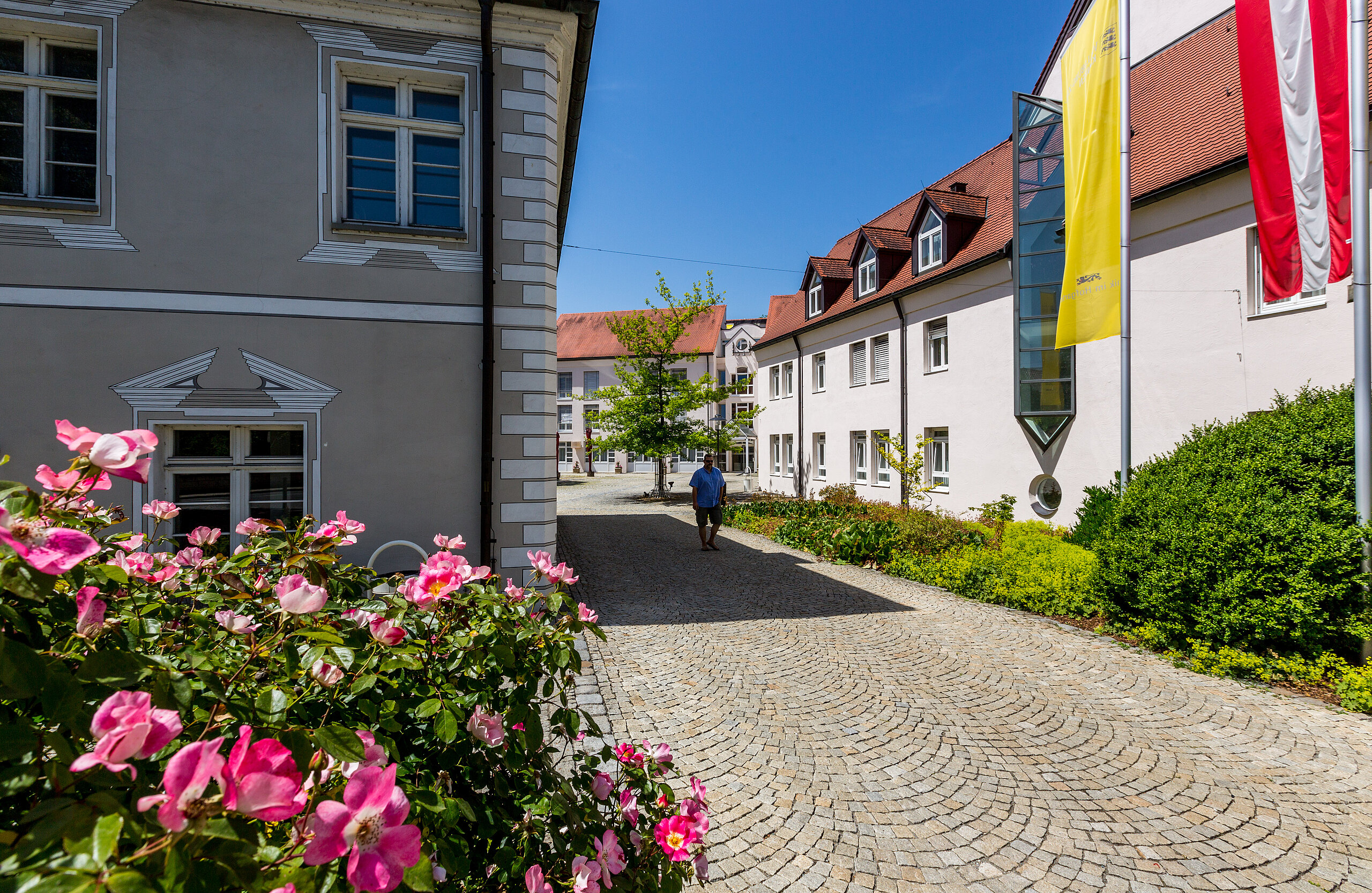 Zugang zur Klinik im Hofgarten mit gepflastertem Weg, Blumenbeeten und Gebäuden mit Fahnen an der Fassade