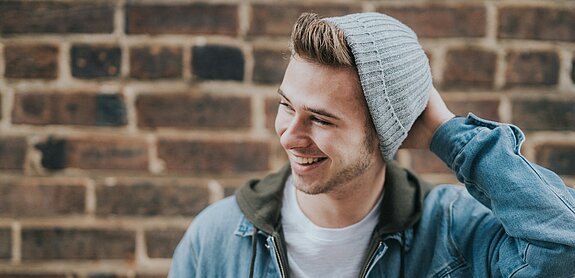 Ein lächelnder Jugendlicher mit Strickmütze vor einer Mauer.