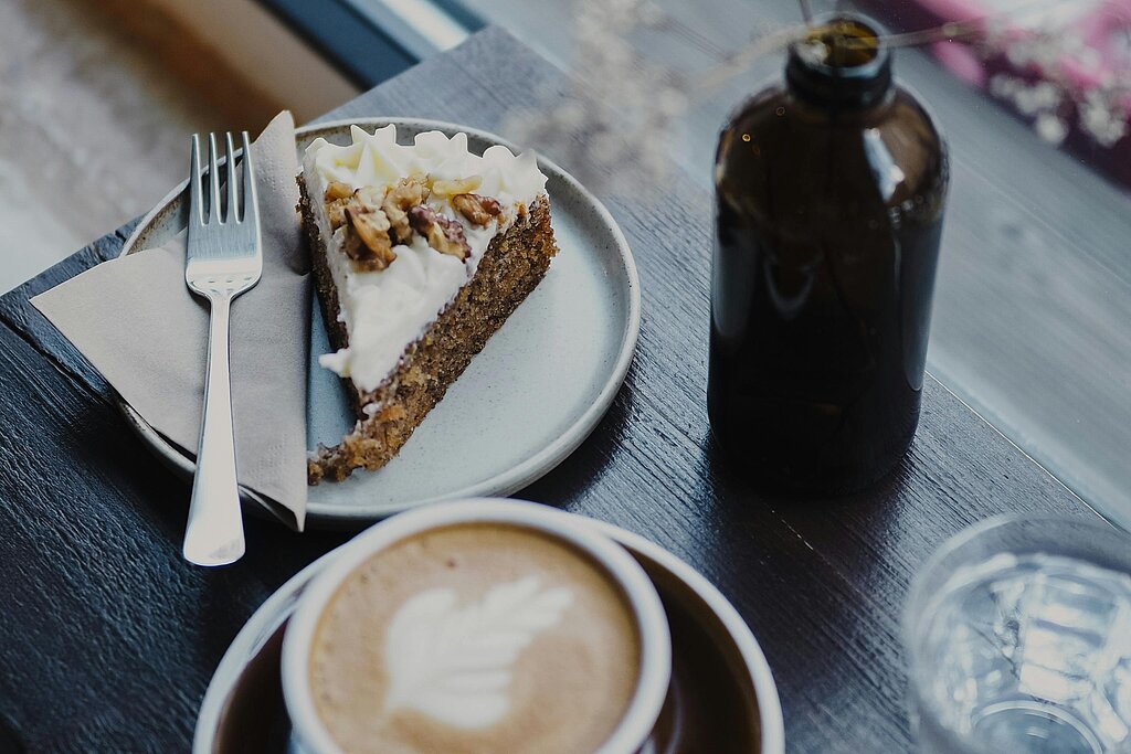 in stilvoll angerichteter Kaffeetisch in einem Café mit einem Cappuccino, einem Stück Kuchen auf einem Teller mit Gabel und Messer, einer kleinen Glasflasche und einem Glas Wasser