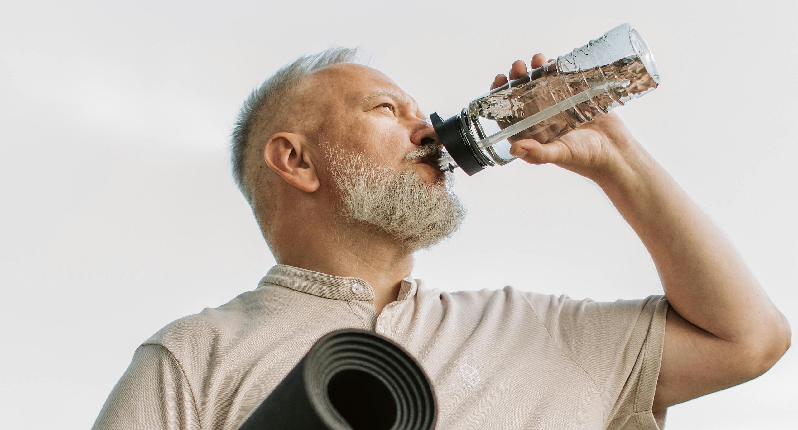 Ein älterer Mann trinkt aus einer Wasserflasche und trägt eine zusammengerollte Gymnastikmatte unter dem Arm.