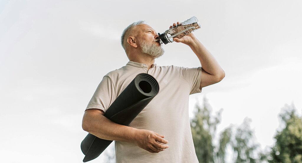 Ein älterer Mann trinkt aus einer Wasserflasche und trägt eine zusammengerollte Gymnastikmatte unter dem Arm.