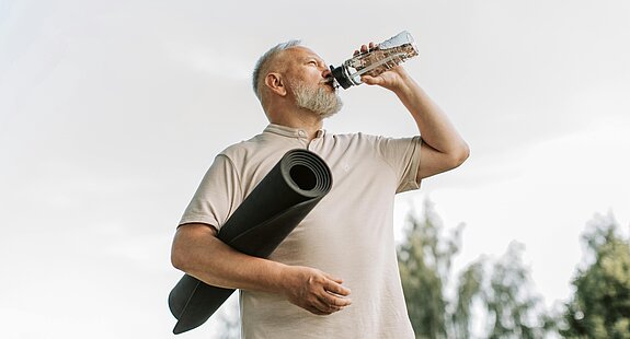Ein älterer Mann trinkt aus einer Wasserflasche und trägt eine zusammengerollte Gymnastikmatte unter dem Arm.