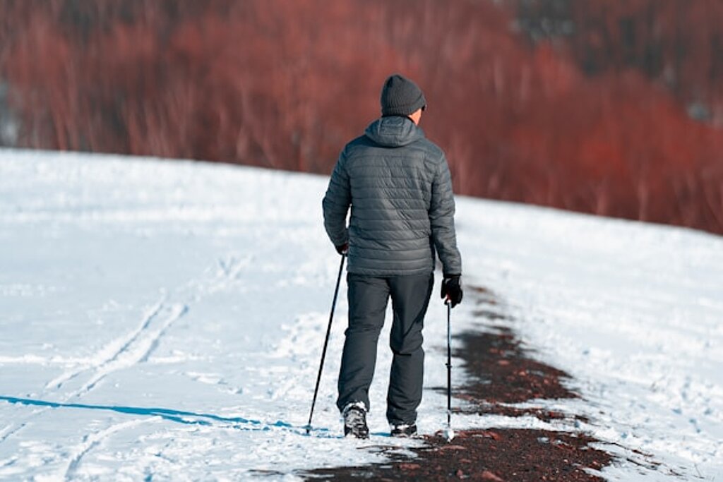 Ein Mann läuft mit Walkingstöcken einen verschneiten Feldweg entlang, im Hintergrund sind rotfarbene Bäume zu erkennen.