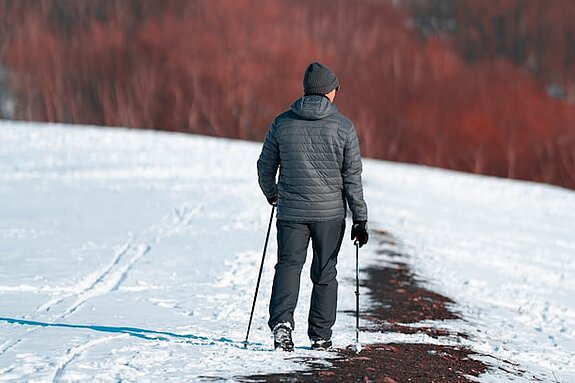 Ein Mann läuft mit Walkingstöcken einen verschneiten Feldweg entlang, im Hintergrund sind rotfarbene Bäume zu erkennen.