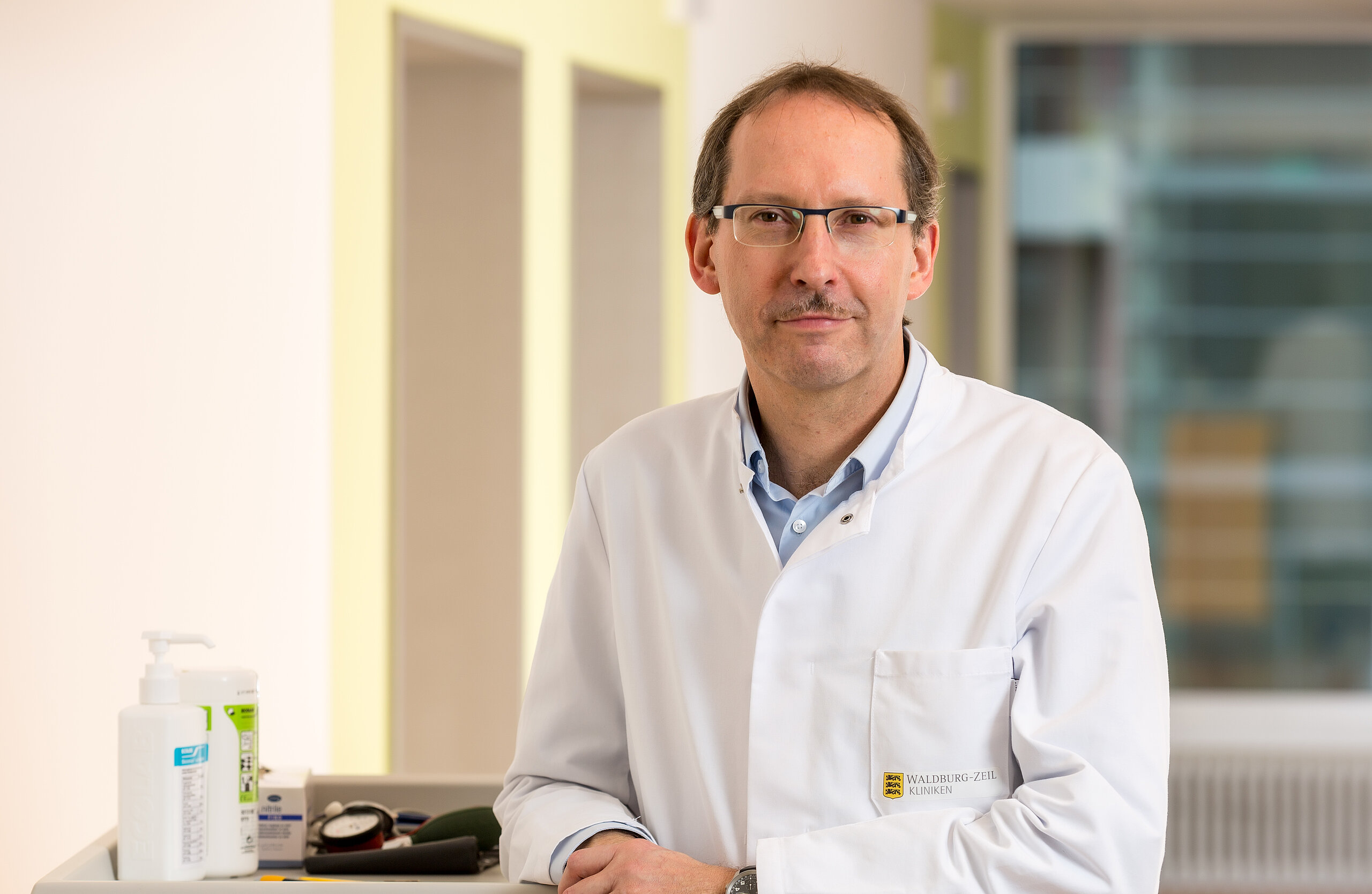 Dr. Klaus Müller, Chefarzt der Klinik im Hofgarten, steht an einem Stationswagen und lächelt freundlich