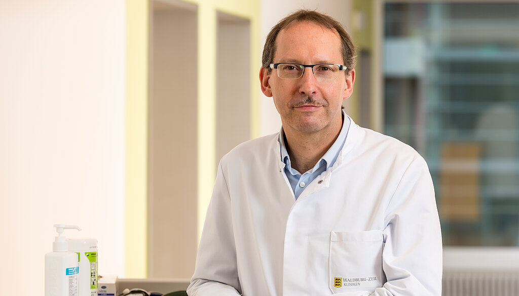 Dr. Klaus Müller, Chefarzt der Klinik im Hofgarten, steht an einem Stationswagen und lächelt freundlich